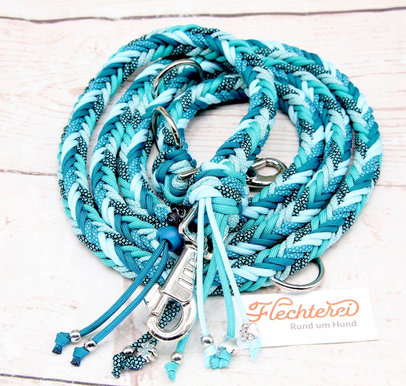 May include: A braided dog leash in shades of blue and turquoise with a silver clip and beads. The leash is made of paracord and has a braided design. The leash is labeled "Flechterei Rund um Hund".