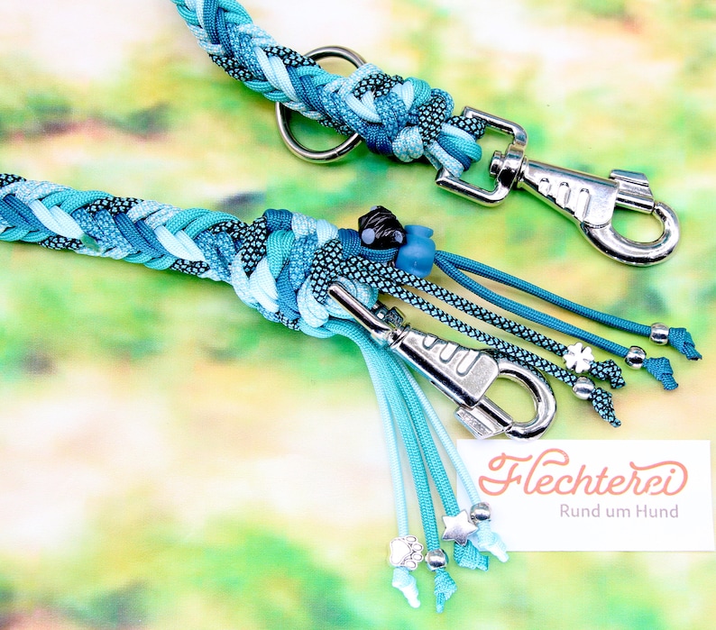 May include: A turquoise and blue braided dog leash with silver hardware and turquoise and blue tassels. The leash has a silver clasp and a silver ring. The leash is decorated with silver beads and stars. The text "Flechterei Rund um Hund" is visible in the image.