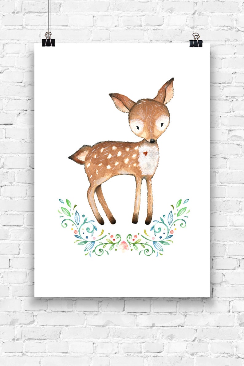 May include: Watercolor illustration of a fawn with brown fur and white spots, standing above a floral wreath. The fawn has large eyes and a small red heart on its chest. The artwork is printed on white paper and displayed on a white brick wall.