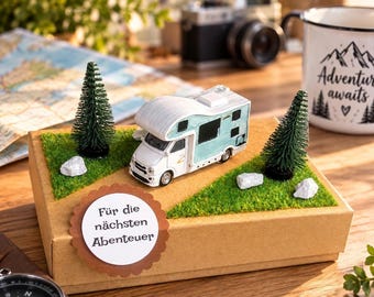 Könnte beinhalten: Eine braune Pappschachtel, dekoriert mit grünem Kunstrasen, zwei Miniatur-Tannenbäumen, weißen Steinen und einem kleinen weiß-hellblauen Wohnmobil-Modell. Ein rundes weißes Etikett auf der Vorderseite trägt den Text "Für die nächsten Abenteuer".