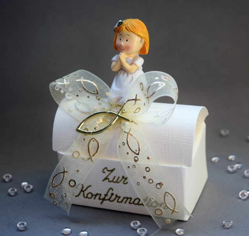 May include: A white gift box with a textured surface and the text Zur Konfirmation in gold script. A translucent ribbon with gold fish patterns and a gold fish charm is tied on top, topped with a small figurine of a child with orange hair.