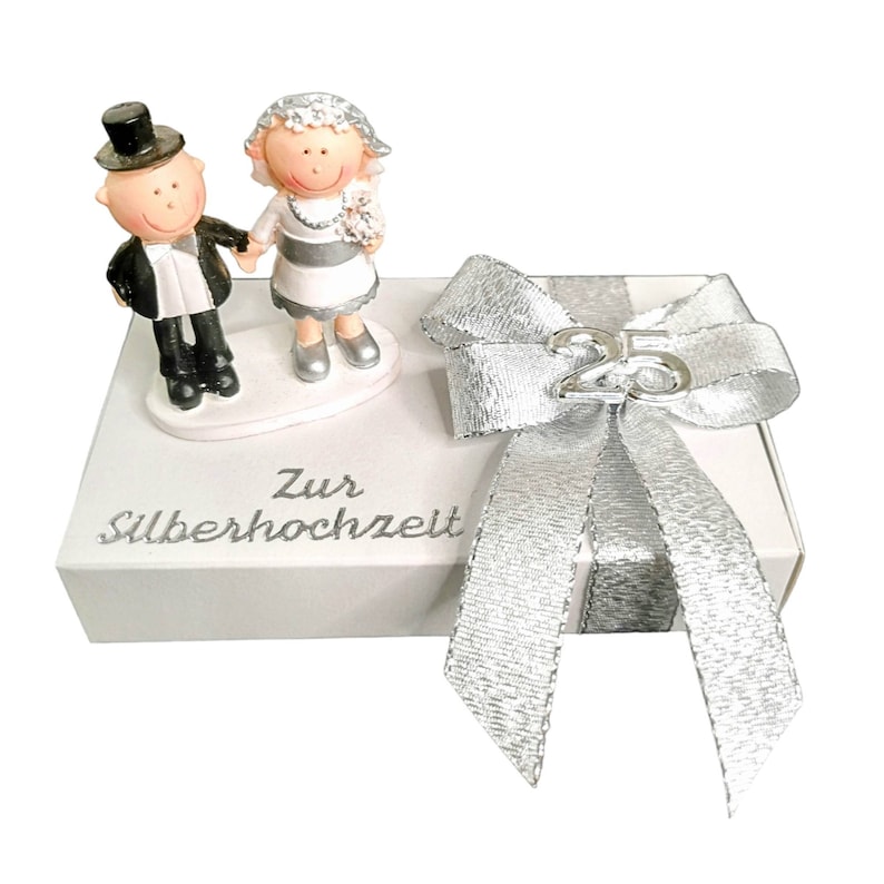 Puede incluir: Una caja de regalo blanca con una cinta y un lazo plateados. La caja tiene las palabras "Zur Silberhochzeit" impresas en la parte delantera. Una peque&ntilde;a figura de una novia y un novio est&aacute; unida a la parte superior de la caja.