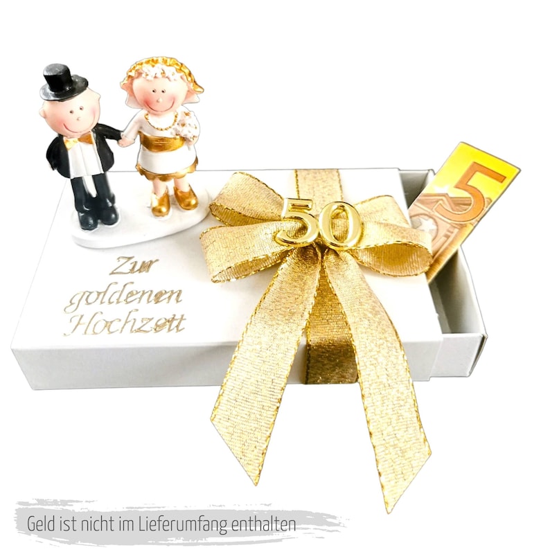 May include: A white gift box with a gold bow and a gold number 50 on top. The box has a white card with the text "Zur goldenen Hochzeit" on it. There are two small figurines of a bride and groom on the box. The box is decorated with a gold ribbon.