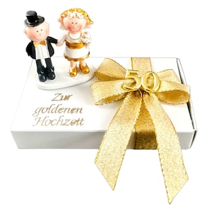 May include: White gift box with a gold ribbon and bow. The bow has a gold number 50 in the center. The box has a small figurine of a bride and groom on top. The box says "Zur goldenen Hochzeit" which translates to "For the golden wedding anniversary".
