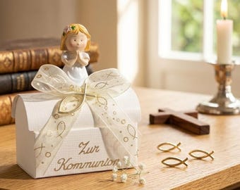 May include: A white keepsake box with a decorative ribbon and a small figurine of a girl with her hands clasped in prayer. The box has the words "Zur Kommunion" in gold script. Gold fish-shaped charms and a wooden cross are also present.