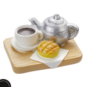Könnte beinhalten: Eine silberne Miniatur-Teekanne, eine weiße Kaffeetasse mit brauner Flüssigkeit und ein gelbes Gebäck auf einem Holztablett.