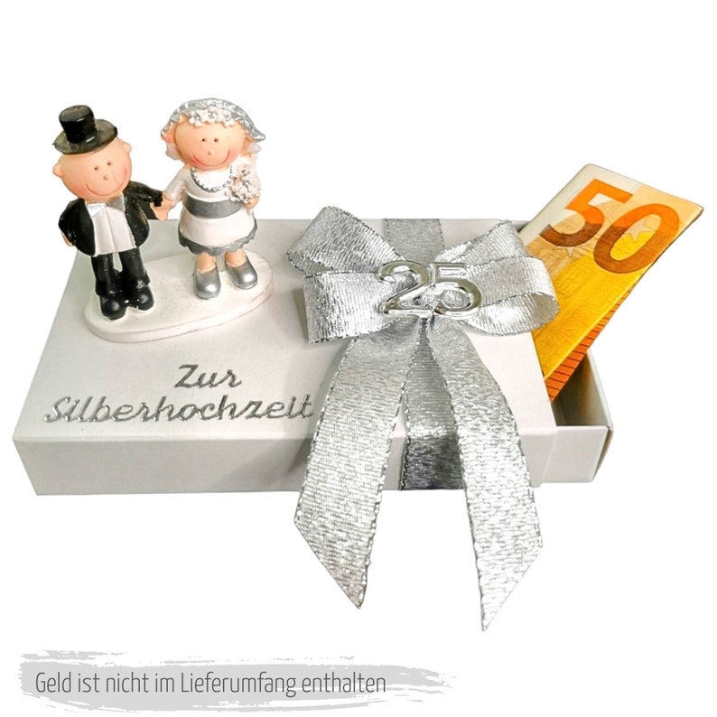 Puede incluir: Una caja de regalo blanca con un lazo plateado y el n&uacute;mero 25 en ella. La caja tiene una inscripci&oacute;n plateada que dice "Zur Silberhochzeit". Hay dos peque&ntilde;as figuritas de una novia y un novio en la parte superior de la caja. La caja est&aacute; abierta y un billete de 50 euros sobresale de ella.