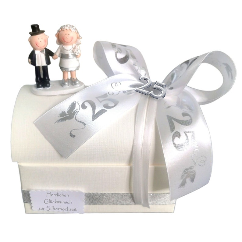 May include: A white gift box with a silver ribbon and a silver charm with the number 25. The box has a silver trim and a small figurine of a bride and groom on top. The box has the text "Herzlichen Gl&uuml;ckwunsch zur Silberhochzeit" on the bottom.
