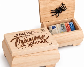 May include: A light-colored wooden box with a hinged lid featuring a black spider attached to the inside. The exterior lid is engraved with the text "EIN PAAR SCHEINE, um Träume zu spinnen" and a small spiderweb. Euro banknotes are inside.