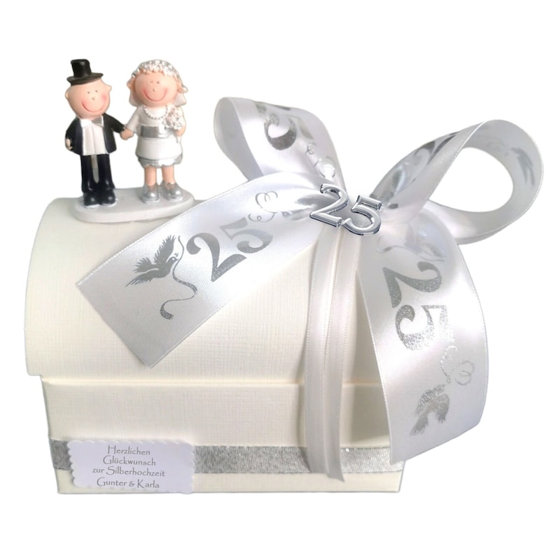 May include: A white gift box with a silver ribbon and a silver 25 charm. The box has a small figurine of a bride and groom on top. The box has a label that says "Herzlichen Gl&uuml;ckwunsch zur Silberhochzeit Gunter & Karla".