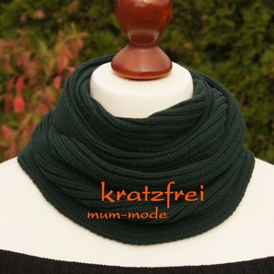 Könnte beinhalten: Dunkelgrüner Strick-Cowl-Schal auf einem Schaufensterkopf. Der gerippte Schal trägt den orangefarbenen Schriftzug "kratzfrei mum-mode". Der Hintergrund ist eine verschwommene Ansicht von grünem und rotem Laub.
