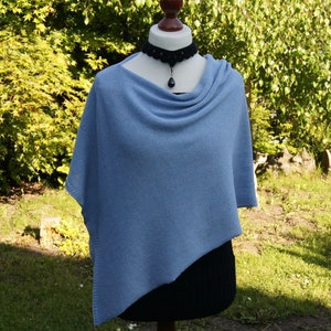 Könnte beinhalten: Hellblauer Strickponcho auf einer Schaufensterpuppe. Der Poncho hat einen drapierten Rollkragen und einen asymmetrischen Saum. Eine schwarze Perlenkette mit Anhänger wird zum Poncho getragen. Die Schaufensterpuppe steht auf einem Holzständer vor grünem Hintergrund.