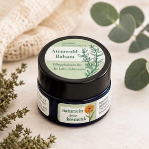 Könnte beinhalten: Ein dunkelblauer Kosmetiktiegel mit schwarzem Deckel und einem Etikett mit der Aufschrift Atemwohl-Balsam, handmade NATURKOSMETIK und Pflegebalsam für die kalte Jahreszeit, sowie einem weiteren Etikett mit naturrein deine kosmetik und einer Blume.