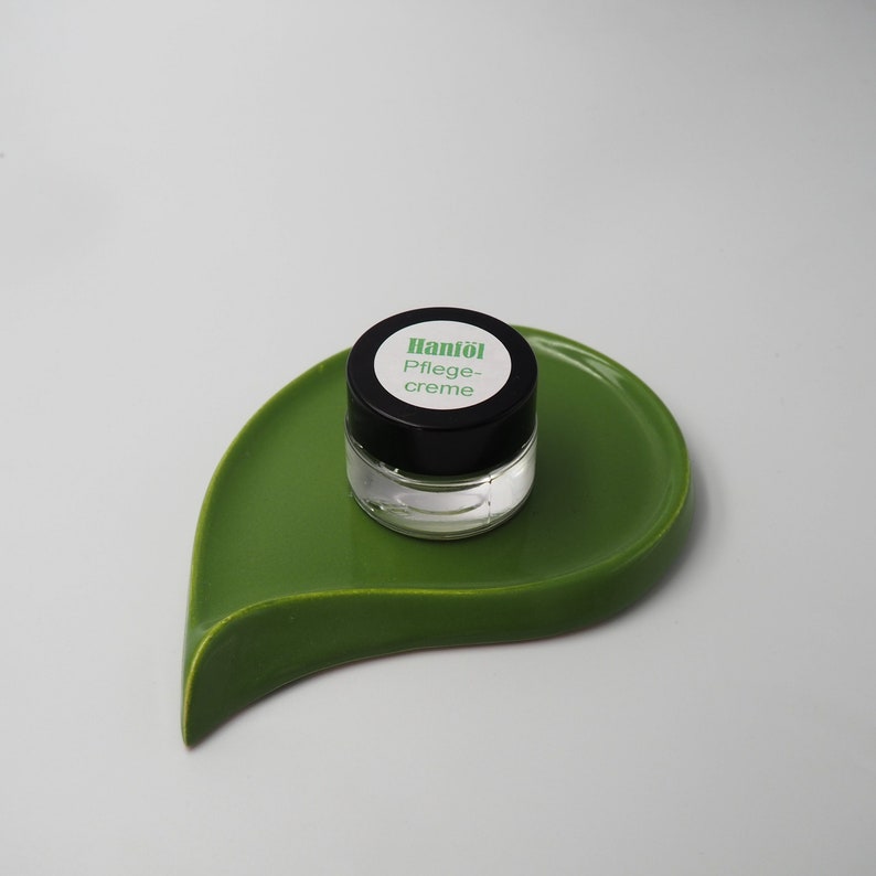 May include: A small, clear glass jar with a black lid and a white label that reads "Hanf&ouml;l Pflege-creme". The jar is sitting on a green leaf-shaped dish.