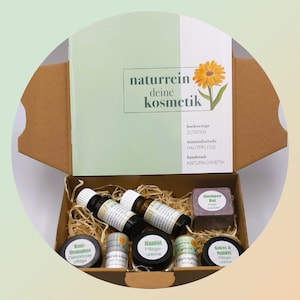Può includere: Una scatola di cartone marrone con un coperchio verde. Il coperchio ha il testo "naturrein deine kosmetik" in bianco. La scatola contiene una varietà di prodotti naturali per la cura della pelle, tra cui una saponetta per capelli, una crema per le mani e oli essenziali. I prodotti sono tutti etichettati in tedesco.