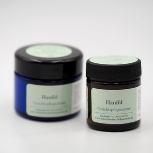 May include: Two small jars of face cream with green labels. The labels read "Hanf&ouml;l Gesichtspflegecreme" and "Hanf&ouml;l Gesichtspflegecreme". The jars are on a white surface.