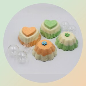 Può includere: Quattro bombe da bagno colorate a forma di dessert, decorate con cuori e fiori. Le bombe da bagno sono nei toni dell'arancione, del verde e del bianco, con sfere di vetro trasparente sparse.