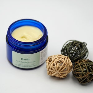 May include: A blue jar of facial cream with the label "Hanf&ouml;l Gesichtspflegecreme". The jar is sitting on a white surface with three decorative balls made of woven material.