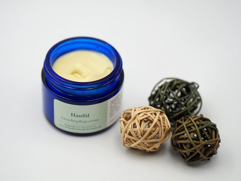 May include: A blue jar of facial cream with the label "Hanf&ouml;l Gesichtspflegecreme". The jar is sitting on a white surface with three decorative balls made of woven material.