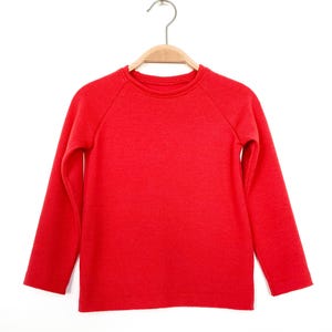 Può includere: Una maglietta rossa brillante a maniche lunghe, esposta su una gruccia di legno. La maglietta ha un girocollo e maniche a raglan. Il colore unito e il design semplice la rendono un capo versatile.
