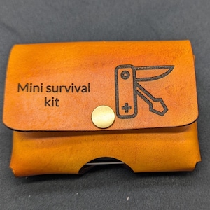 May include: A brown leather case with a brass button closure. The case is engraved with the text "Mini survival kit" and a graphic of a multi-tool.