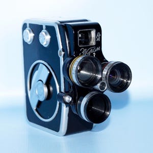 Puede incluir: Una cámara de cine vintage negra y plateada con múltiples lentes y diales. La cámara tiene el texto "Miram 3" en el lateral. La cámara está sobre un fondo azul claro.