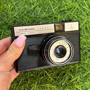 May include: A black and silver vintage Smena Symbol camera with a lens and text. The camera is held against a background of green grass. The camera has a silver top and a black body with a lens.