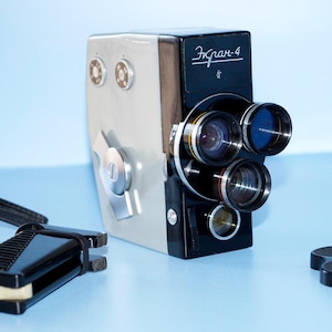 Puede incluir: Cámara de cine Ekran-4 vintage en blanco y negro con múltiples lentes. La cámara tiene una correa negra y un accesorio negro. El texto "Ekran-4" es visible en la cámara.