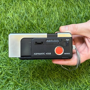 Op de afbeelding: Een Agfamatic 4008 sensor camera met een zwarte en zilverkleurige rechthoekige behuizing. Het apparaat is voorzien van een opvallende rode ronde ontspanknop, een afstandsschaal in meters, een tele-schakelaar en een zilveren metalen polsbandje.