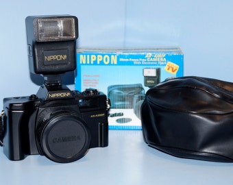 May include: A black 35mm film camera with a built-in flash and a black leather camera bag. The camera is labeled "NIPPON" and the box is labeled "NIPPON 35-2000 Zoom Focus Free CAMERA with Electronic Flash TV".