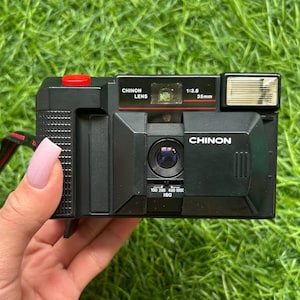 以下が含まれることがあります： 赤色のシャッターボタンと内蔵フラッシュを備えた黒色のChinon 35mmフィルムカメラ。カメラにはChinonレンズとISO設定があります。カメラは緑色の草の背景に対して保持されています。
