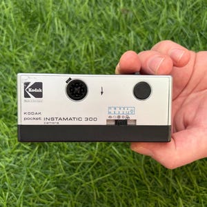 May include: A silver and black Kodak pocket Instamatic 300 camera. The front features the Kodak logo, a circular lens opening, a black shutter button, and a sliding exposure control panel with weather icons and distance markings in meters.