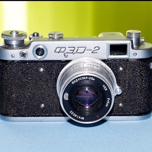 Puede incluir: Una cámara vintage plateada y negra con una lente prominente. El cuerpo de la cámara tiene un acabado negro texturizado, con detalles y diales plateados. La lente está etiquetada como "INDUSTAR-26m" y "f=50mm 1:2.8". La cámara está sobre una superficie azul.