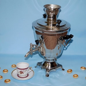May include: A silver samovar with a black handle and a spout, sitting on a light blue surface with a white teacup and saucer and several small, round, brown pastries.