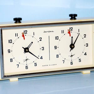 May include: Cream-colored vintage chess clock with two clock faces. Each face has black numerals, black hands, and a red second hand. The clock has a silver frame and black knobs. The text "Jantas" and "Made in USSR" are visible.
