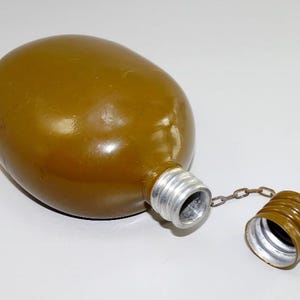 May include: An olive-drab, oval-shaped canteen with a silver metal neck and a matching cap attached by a chain. The cap is unscrewed and lying next to the canteen. The canteen is a military-style water bottle.