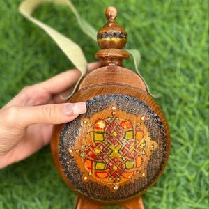 May include: A wooden flask with a decorative design. The flask is brown with a detailed, colorful pattern on the front, featuring red, orange, and yellow accents. It has a small handle and a decorative top.