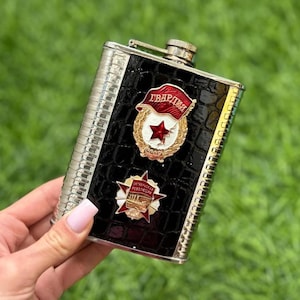May include: A stainless steel flask with a black, textured front panel featuring two red and gold emblems. The flask has a silver cap and trim, and is held against a backdrop of green grass.