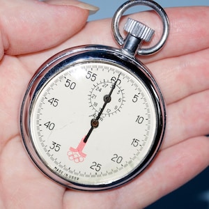 Op de afbeelding: Een vintage stopwatch met een zilveren behuizing en een witte wijzerplaat, vastgehouden in een hand. De wijzerplaat toont cijfers en markeringen voor het meten van tijd, met een rood en roze Olympisch symbool. Het is een klassiek tijdmeetapparaat.