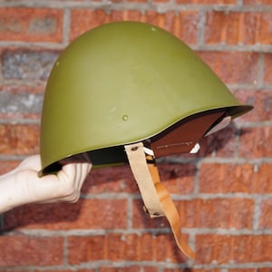 May include: A green military helmet with a brown leather strap. The helmet is held in a hand against a brick wall background.
