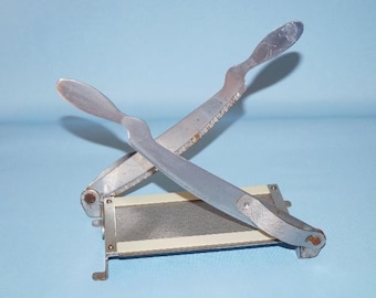May include: A vintage paper cutter with a silver metal frame and blades. The blades are crossed, with one serrated edge. The base has a white stripe. The background is a light blue.