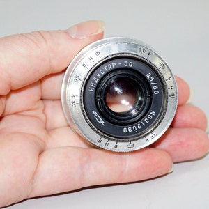 Op de afbeelding: Een zilveren camera lens met zwarte markeringen. De lens heeft een zwarte diafragmaring met markeringen voor f/3.5, f/4, f/5.6, f/8, f/11 en f/16. De lens heeft ook een zwarte scherpstelring met markeringen voor 3.5, 4, 5.6, 8, 11, 16 en 32. De lens is gemarkeerd met "INDUSTAR-50" en "N6312099".