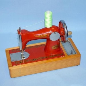 May include: A vintage red metal sewing machine mounted on a rectangular wooden base. The machine features gold floral detailing, a silver hand wheel, and a spool of light green thread positioned on top. The needle plate is silver metal.