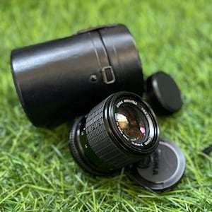 May include: A black Sigma Mini-Tele camera lens with a lens cap and a black leather case. The lens is lying on green grass. The lens has the text "SIGMA MINI-TELE" and "1:4 f=135mm" printed on it.