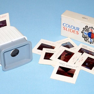 Puede incluir: Un visor de diapositivas de plástico blanco y gris con una diapositiva insertada. El visor está al lado de una caja de diapositivas de color etiquetada como "COLOUR SLIDES" y una pila de diapositivas de color con imágenes de varios lugares.