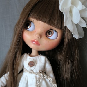 Könnte beinhalten: Puppe mit braunem Haar, Pony und großen braunen Augen. Die Puppe hat Sommersprossen und trägt ein weißes Kleid mit einem Holzknopf und einem dekorativen Gürtel. Eine große weiße Blume schmückt das Haar der Puppe.