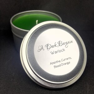 May include: A silver-colored candle tin with a green candle inside and the lid slightly ajar. The lid features the text "A Dark Bargain Warlock" and lists the scents: Absinthe, Currants, Blood Orange.