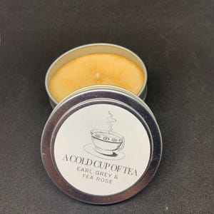 May include: A silver tin candle with a white label that reads "A Cold Cup of Tea Earl Grey & Tea Rose". The label features a black and white illustration of a teacup with steam rising from it.