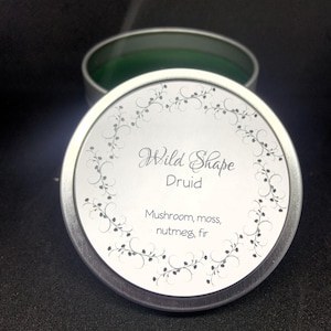 May include: A silver-colored round tin with the text "Wild Shape Druid" and the scent notes "Mushroom, moss, nutmeg, fir." The tin is partially open, revealing a green interior. A decorative floral border encircles the text.