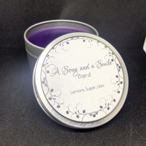 May include: A Song and a Smile Bard candle in a silver tin. The candle has a purple wax and the lid has the text "A Song and a Smile Bard" with the scent notes "Lemons, Sugar, Lilies". The lid is decorated with a floral design.